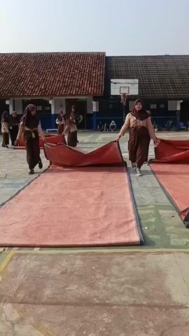 The video shows a group of young people, likely students, in a schoolyard. They are wearing brown uniforms with hijabs. The students are in the process of unfolding and laying out large, red tarps on the paved ground. Some students are holding the edges of the tarps and pulling them flat, while others are already standing near the laid-out sections. In the background, there is a building with a tiled roof and a basketball hoop. The lighting suggests it is daytime. One student on the right is holding a red bottle.