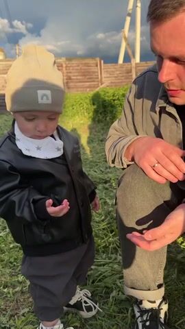 A toddler in a beige beanie with a rainbow emblem, a black leather jacket, gray sweatpants, and white tennis shoes stands on a grassy lawn and holds out their hand. Perched on their index finger is a ladybug. An adult with dark hair and a gray jacket kneels next to the toddler, observing. A wooden fence and power lines are visible in the background beneath a cloudy sky.