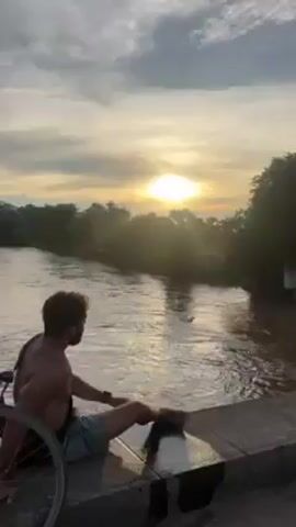The video shows a man sitting on the edge of a bridge, overlooking a river. The sun is setting behind him, creating a golden glow on the water. The river is wide and the current is strong. There are trees and vegetation lining the riverbanks, with hills or mountains visible in the distance. A bicycle is parked near the man. The man is shirtless and wearing shorts. The sky is blue with some clouds.