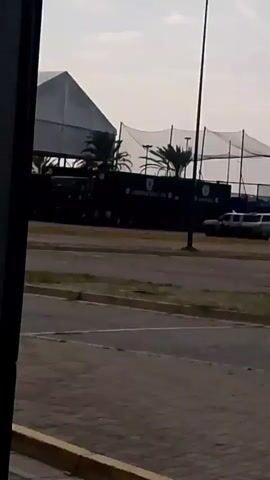 This is a vertical, shaky video, likely recorded from inside a moving vehicle. The view is angled towards the right, showing a paved area with yellow curbs and a bit of greenery. In the background, a row of dark-colored trucks is visible, possibly parked. Further back, there's a large white structure with a pitched roof and some smaller structures. A sign with some text is also partially visible. A smaller vehicle, possibly an ambulance or a modified van, is also present. The overall impression is of an outdoor, possibly commercial or industrial area, with vehicles and buildings. The lighting suggests it is daytime, but the sky is overcast.