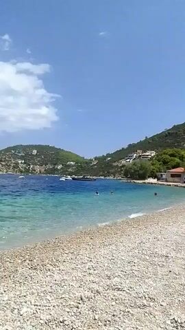 The video shows a panoramic view of a pebble beach with clear turquoise water lapping at the shore. In the distance, a large body of water extends to the horizon, dotted with several boats. Green, hilly terrain covered with houses and trees rises from the water's edge on the right side of the frame. The sky is blue with scattered white clouds. A few people are visible swimming or wading in the shallow water. The camera is stationary.