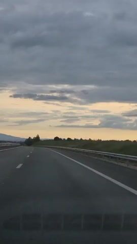 The video shows a view from inside a car driving on a highway. The highway has multiple lanes and is separated by a barrier. The sky is cloudy and appears to be overcast with some lighter patches of sky visible. There are trees and vegetation on the sides of the road. In the distance, there are some hills or mountains. The video captures the motion of the car moving forward on the highway.