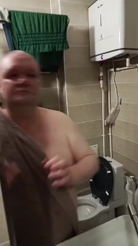 A man with a shaved head and a towel draped over his shoulder is in a bathroom. He is looking into a mirror and touching his nose with his fingers. He then makes a kissing gesture with his lips and smiles at the mirror. The bathroom has tiled walls, a towel rack with towels, a white electric water heater on the wall, and what appears to be a sink.