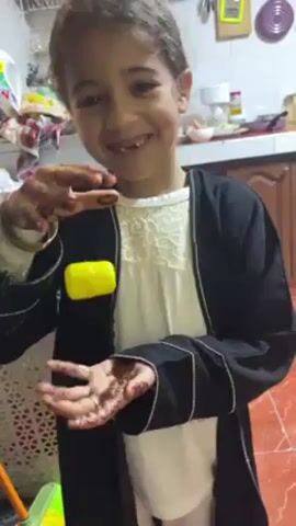 A young child with missing front teeth is standing in what appears to be a kitchen. The child has henna on their hands and is wearing a black robe over a white outfit, along with a bracelet. The child is smiling and looking at the camera while appearing to levitate a yellow object between their hands. The kitchen background includes various household items, cabinets, and red tile flooring. The child speaks during the video.