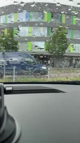 The video captures a view from inside a vehicle looking out through a rain-splashed windshield on a seemingly overcast day. Numerous large raindrops are visible on the glass, indicating heavy rain.

In the background, a modern building with distinctive green and blue facade panels stands behind some trees. The scene appears to be a street or parking area in front of this building, possibly in Waldshut, Germany, as per the provided context.

During the short clip:
- At the 0:00 mark, a dark blue van with a blurry red circular logo on its side drives past from left to right.
- After the van, a white car also drives past in the same direction, visible around the 0:02-0:03 mark.

The foreground of the video shows the car's dashboard, including a speaker grille and a portion of a black suction cup mount on the left side. No specific individuals are clearly visible, and no discernible text can be read on the passing vehicles or the building.