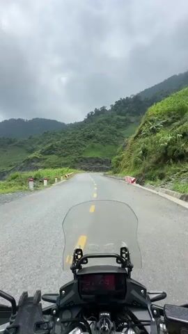 The video captures a scenic motorcycle ride through a mountainous region, likely Minh Sơn Commune, Vietnam.

The opening shot provides a first-person perspective from the motorcycle, showing a winding paved road with a yellow double solid line down the center. Lush green, cloud-covered mountains flank the road, suggesting a remote, hilly landscape. The motorcycle's dashboard is visible at the bottom of the frame, displaying "44" (likely speed in km/h) and a "N" indicator, along with other unreadable digital information.

As the camera pans, red and white striped concrete barriers are seen lining the edges of the road, particularly on curves overlooking valleys. The scene transitions to wider shots revealing a picturesque valley with extensive green foliage, possibly agricultural crops like corn, under a gradually clearing sky. The weather evolves from an overcast and hazy sky to one with visible patches of blue and brighter sunlight, illuminating the rugged landscape and distant misty peaks.

Mid-video, a selfie-style shot captures two individuals on the motorcycle. The rider in front wears a red jacket, sunglasses, and a bright yellow full-face helmet with a GoPro camera mounted on the chin. The passenger behind is dressed in a grey jacket, sunglasses, and a grey full-face helmet, actively pointing towards the scenic vista to their right. The winding mountain road, bordered by steep, verdant slopes and offering glimpses of distant peaks, is central to the journey. The ride appears to be a spirited exploration of a beautiful, undulating landscape under dynamic weather conditions.