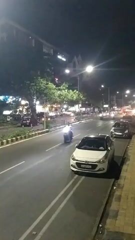 The video shows a nighttime street scene with moderate traffic. A white car is parked on the right side of the street, next to a tiled sidewalk. A scooter with a bright headlight is approaching in the lane closest to the camera. Other cars and motorcycles are visible in the distance, along with streetlights illuminating the road.  Trees line the median and the opposite side of the street. Buildings are visible in the background.