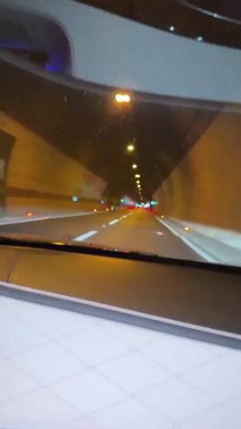 A first-person perspective video shows a car driving through a well-lit tunnel. The camera is positioned behind the windshield, looking forward. The tunnel walls are visible, lined with yellow lights at intervals. White lane markings are clear on the road ahead. Red and green lights from other vehicles are seen in the distance, creating a bokeh effect. The interior of the car, including part of the dashboard and a diamond-stitched seat, is visible in the foreground. The overall lighting suggests it is either nighttime or the tunnel is dimly lit.