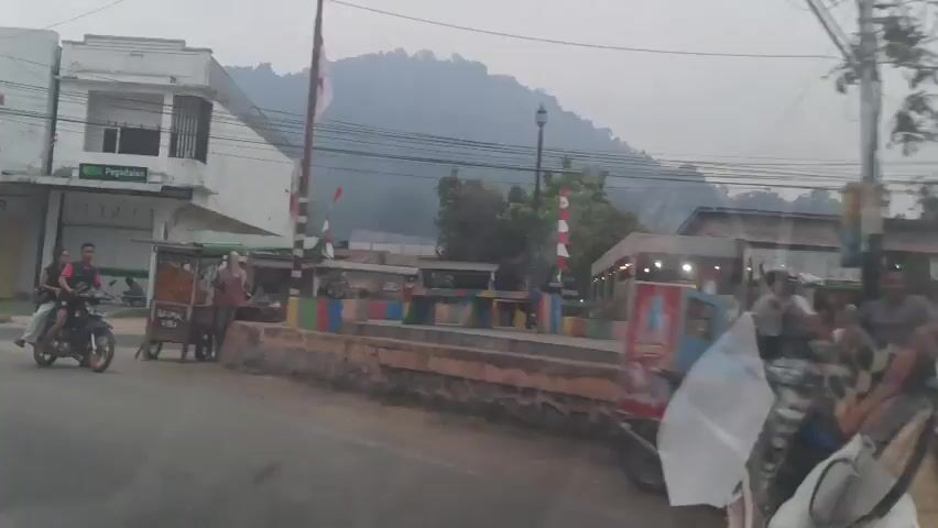 The video shows a street scene, likely in Southeast Asia, with several buildings lining the road. On the left, a white building with a red Honda logo is visible, appearing to be a motorcycle dealership. Next to it is another white building with text that appears to be "Pegadaian," a type of pawnshop in Indonesia. A street food vendor's cart is set up in front of this building. Several motorcycles are parked in front of the businesses. In the foreground, a person is riding a motorcycle with two passengers. Further down the street, there is a colorful, low wall with flags. A large hill or mountain is visible in the background, partially obscured by mist or haze. The sky is overcast, suggesting it might be early morning or late afternoon. The overall atmosphere is that of a typical commercial street in a developing urban or semi-urban area.