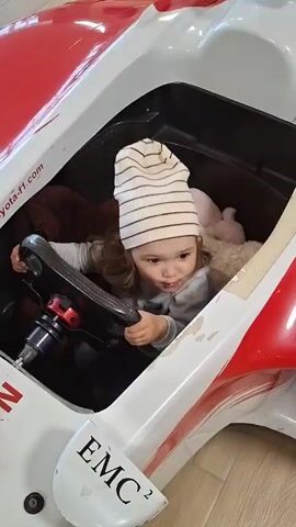 A toddler wearing a striped beanie sits in the driver's seat of a toy race car. The car is white with red accents and has the text "EMC²" visible on its side. The child holds the steering wheel, appearing focused. The setting appears to be indoors, possibly a waiting area or play space, with chairs visible in the background. The lighting suggests it's daytime. No specific time of day or weather conditions can be determined. The child's expression is neutral, with a slight furrow of the brow. The toy car itself seems to be a replica of a Formula 1 car, with text like "toyota-f1.com" and "DENSO" visible on its body. There are also miniature toy cars placed on the front of the race car.