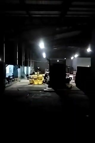The video pans across a dimly lit warehouse. Several rows of overhead lights illuminate the space. In the foreground, the camera is positioned low, looking across a dark floor. To the left, a collection of yellow barrels or containers are stacked. Further back, the warehouse appears to be filled with various objects, some of which are obscured by shadow. There are indistinct figures of people in the background, moving around. The overall impression is a busy, industrial environment, possibly a storage or distribution center, captured during low-light conditions.