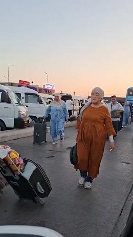 The video shows a group of people with luggage at what appears to be a transportation hub or a parking area. In the foreground, an older woman in an orange dress and headscarf is walking away from the camera, pulling a black bag. To her left, another woman in a blue patterned dress and headscarf is pulling a rolling suitcase. Behind them, a blue bus is visible, as are several white vans and cars. Other people are seen walking with luggage in the background. The sky is a soft orange and pink, suggesting it is either sunrise or sunset. Some of the vehicles have license plates visible. The ground is asphalt.