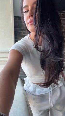 A woman stands on a balcony in front of a brick wall and a white railing. She is wearing a white crop top and grey sweatpants with a frilly drawstring. She has long, dark hair which she runs her hand through.