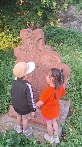 Two young children, a boy and a girl, are standing in front of a large, intricately carved stone cross. The boy, wearing a beige baseball cap and a black jacket with white trim, points at the cross. The girl, with braided hair and wearing an orange t-shirt, also touches the cross. They are in an outdoor setting with lush green foliage and grass surrounding them. The sun appears to be setting, casting a warm light on the scene.