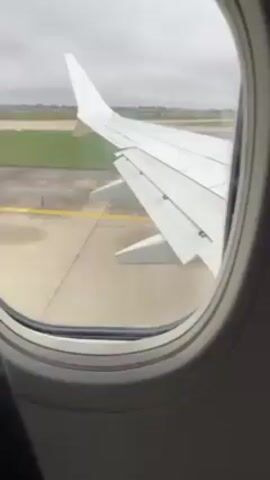 The view from an airplane window shows a white wing of the aircraft on a wet tarmac with yellow lines. The weather outside is overcast. Distant buildings are visible in the background. The airplane is moving slowly.