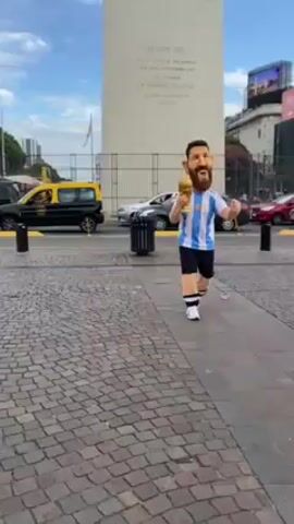 The video shows a person in an Argentina soccer jersey and a Lionel Messi mascot head, holding the World Cup trophy, walking in front of a tall monument on a brick-paved plaza. The person in the Messi costume waves to a couple and a young girl who approaches him, then continues walking and waving in front of the monument. Cars and buildings are visible in the background. The sky is partly cloudy.