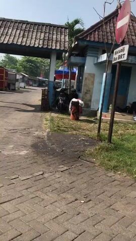 The video opens on a brick-paved walkway leading towards an arched gateway. Beyond the gateway, the scene opens up to a bustling area with numerous market stalls and a building with a blue and white facade. A motorcycle is parked near the building, and a person is seated in the background. A traffic sign is visible on a pole, indicating "KECUALI BUS BRT" (Except BRT Bus). The sky is clear and the sun is shining, casting shadows on the ground. The environment appears to be an outdoor marketplace or transportation hub.