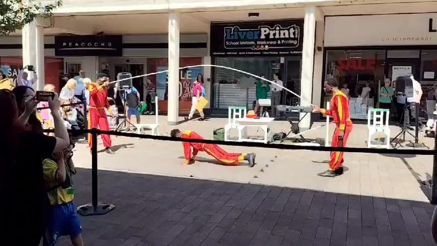 The video shows an outdoor street performance in front of shops. Three individuals dressed in red and yellow outfits are engaged in a rope skipping activity. One person is on the ground in a push-up position, while two others stand and swing a long white rope. A fourth person in a similar outfit stands to the right, holding a rope. Several onlookers are watching the performance. In the foreground, a woman is holding a phone to record, and a young boy is covering his ears. The shops visible include "Peacocks" and "LiverPrint." The ground is paved with brick-like stones, and there are white chairs and a small table set up as part of the performance area. The scene appears to be taking place during the daytime, with sunlight illuminating the area.