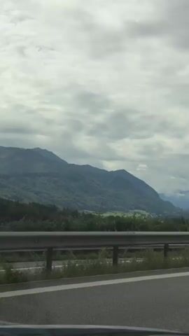 The video shows a view from a vehicle moving along a highway. The highway has a gray asphalt surface with a white lane marker. A metal guardrail runs along the side of the highway. Beyond the guardrail is a grassy area with some vegetation. In the distance, there are mountains covered in trees under a cloudy sky. The mountains appear to be a deep green color. The overall impression is of a scenic drive through a mountainous region on a cloudy day.