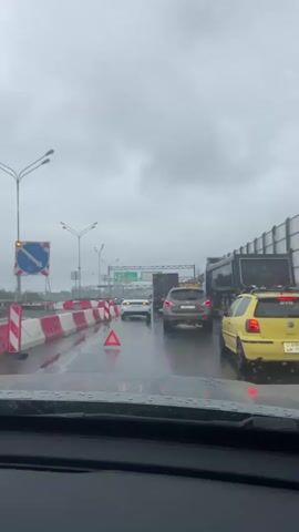 The video shows a traffic jam on a rainy day, likely on a highway given the presence of multiple lanes, overhead signs, and traffic barriers. The road is wet, indicating recent or ongoing rain, and the sky is overcast with thick clouds, suggesting gloomy weather and possibly early morning or late afternoon.

The main subjects are vehicles: a yellow car, a white sedan, a darker SUV, and several trucks. The cars are lined up, indicating slow or stopped traffic. A red triangular warning sign is visible on the road, suggesting a hazard or breakdown ahead. Traffic barriers, painted red and white, are set up along the side of the road, possibly indicating construction or lane closures.

Overhead signs are visible, with green text suggesting directions or destinations. One sign, partially obscured, appears to have Russian text. The license plates on some vehicles are also visible, with characters that appear to be Cyrillic, consistent with the provided location context of Lisy Nos, Russia.

There are no people clearly visible, but the context implies the presence of drivers within the vehicles. The overall emotion conveyed by the scene is one of frustration or tedium due to the traffic congestion and poor weather. The setting appears to be an urban or suburban highway environment.