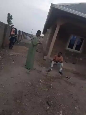 The video shows a dirt yard in a residential area. A woman in a hijab stands near a wall, while a man in a green robe stands facing away from the camera. Another man is sitting on the ground near a building. The man in the green robe raises his hand.
