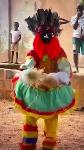 The video shows two people in colorful costumes dancing on a dirt ground. Behind them, a group of children is watching. The dancers have on full face masks with colorful clothing, including feather headpieces. The backdrop of the video includes old, unpainted buildings. The light outside appears bright and sunny.