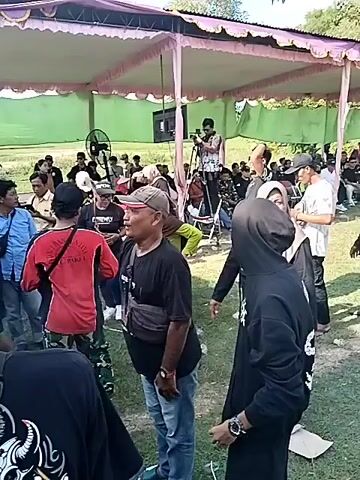 The video depicts a live music performance at an outdoor event. 

The initial scene shows a crowd gathered under a tent-like structure, with people dressed casually. The environment appears to be a field with greenery in the background. A cameraman is filming the event from a tripod.

The video transitions to a shot of a stage with a banner displaying a logo, as well as some musicians. 

The band is performing on stage. There is a male and female vocalist. Other musicians are playing instruments, including drums, guitar, and keyboard. The crowd is enjoying the music. 

Finally, the video shows the back of the stage, with the speaker boxes. People are milling around the equipment. The environment appears to be rural, with trees and a rice field visible in the background.