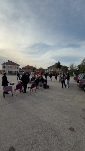 The video shows a group of people gathered in a town square or open paved area with houses surrounding it. In the center of the square, a small brass band of older men in suits are seated on purple chairs and playing instruments including trumpets, trombones, and a tuba. A man stands near the band, possibly conducting or leading. 
Several people are standing or walking around the square. Some people are standing in a small group near the band, watching them play. Others are walking around, passing through the area. In the background, there are red market stalls set up, indicating a possible market or event taking place. The sky is overcast with scattered clouds, suggesting it is late afternoon or early evening.