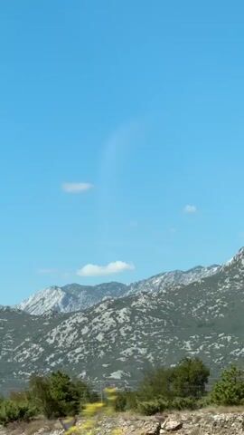 The video shows a view of a mountain landscape under a clear blue sky with a few scattered clouds. A tall, thin lamppost is visible in the foreground, slightly to the left of the center. Beyond the lamppost, the terrain is covered with sparse vegetation, including bushes and some trees. The middle ground is dominated by a rugged mountain range with visible rock formations and patches of greenery. The mountains appear dry and somewhat barren in places. The overall environment suggests a warm, sunny day, possibly in a Mediterranean or similar climate. The camera appears to be moving, as evidenced by the slight blurring of the foreground vegetation and the way the landscape shifts. There are no people visible in the video.