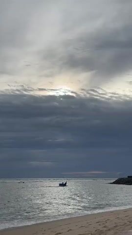 A small blue boat is on the water with a person visible on it. The sky is cloudy with the sun peeking through. There is a beach in the foreground with sand and some debris. To the right of the frame, there is a rocky structure extending into the water.