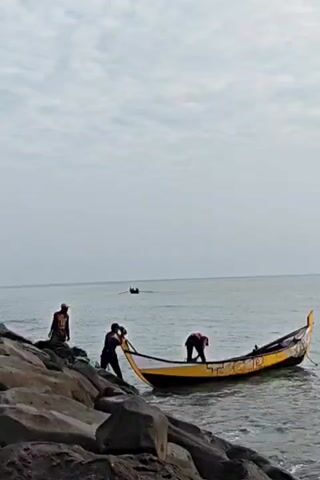 The video shows three people with a boat at the water's edge, next to a rocky bank. The boat is primarily yellow and black. One person stands on the rocks near the boat, another is inside the boat, and a third stands on the rocks further away. In the background, another boat is visible further out on the water. The sky is overcast.