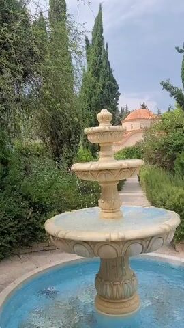A three-tiered fountain with a light beige finish and a blue basin is featured in the foreground. Water is cascading from the upper tiers to the lower ones. The fountain is situated in a garden with lush green foliage and tall, slender trees. In the background, a pathway leads towards a building with a terracotta-tiled roof. The sky is overcast.