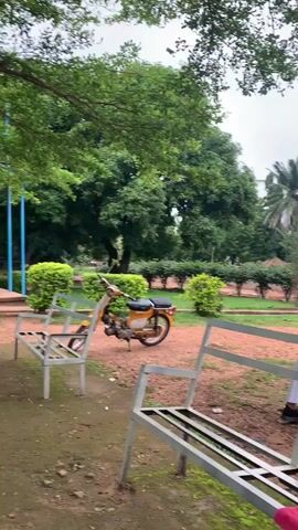 The video opens with a shaky, slightly out-of-focus shot of a park-like setting. A person in white clothing is visible, walking away from the camera. There are white benches and a motorcycle in the background, surrounded by greenery. The scene then shifts to a wider outdoor view. A dirt path leads towards a blue and white building with a gate. The sky is overcast. A ditch runs along the foreground. Trees are scattered throughout the landscape. Several small structures are visible in the distance. The camera pans and zooms slightly, revealing more of the environment, including what appears to be a security booth at the entrance of the building. The video ends with a clear shot of the building's entrance.