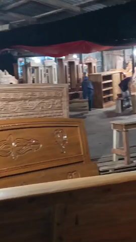 The video shows an outdoor furniture store at night, filled with unfinished wooden furniture including bed frames, benches, tables, and cabinets. Several men are milling about, presumably workers or customers. The area is covered by a metal awning with some red tarps overhead. The lighting is dim and seems to be coming from overhead bulbs.