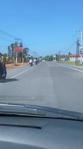 This video captures a view from a vehicle driving on a paved road under a clear blue sky. A line of motorcycles is traveling ahead in the same direction, appearing to be in a procession or a group ride. The motorcycles are spaced out along the road. On the left side of the road, there are utility poles with many wires, a small building with a red sign, and some vegetation including palm trees and other greenery. On the right side of the road, there are more utility poles, a building with a red roof, and houses in the distance, suggesting a somewhat rural or suburban environment. The road itself has yellow lane markings. The perspective is from the driver's or passenger's seat, looking through the windshield, with the dashboard visible in the foreground. The overall impression is of a sunny day and normal traffic on a road in a tropical or subtropical region.