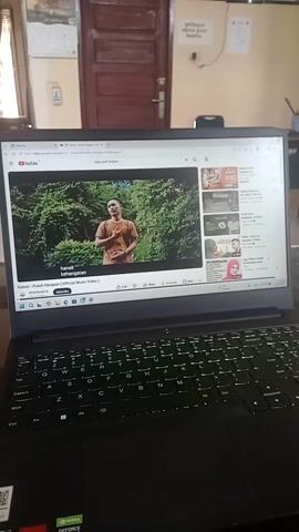 The video displays a laptop screen showing a YouTube page. The video playing on the screen is a music video with a man in a maroon shirt standing in front of a dark background. In the sidebar of the YouTube page, there are thumbnails of other videos, some of which feature people. The laptop itself is black with a backlit keyboard. The scene is viewed from an angle, with the laptop placed on a desk. The background shows a room with a wooden door and a calendar. The lighting in the room appears to be ambient, with some artificial light coming from the laptop screen and keyboard.