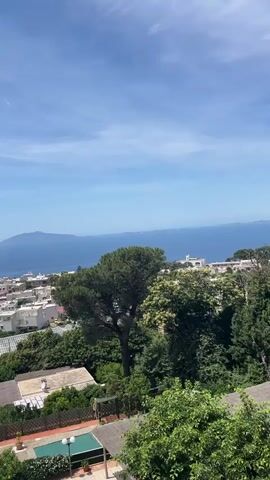 The video pans across a coastal town with white buildings and lush green trees under a bright blue sky with scattered white clouds. In the distance, the deep blue sea stretches out to meet the horizon, where a faint landmass is visible. A small rectangular swimming pool with turquoise water and lounge chairs is situated in the foreground, near a paved patio area with a white umbrella. The town appears to be nestled on a gentle slope, with more buildings and foliage extending upwards towards the sea. The atmosphere suggests a sunny, pleasant day in a Mediterranean setting.