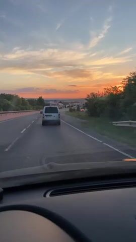 The video shows a view from inside a car driving on a highway towards a sunset. A white van is driving ahead of the car on the highway. The sky is filled with vibrant orange and yellow hues from the setting sun, with some scattered clouds. The highway has multiple lanes and is bordered by trees and vegetation. In the distance, there appear to be some buildings and industrial structures. The interior of the car is visible in the foreground, including the dashboard and part of the steering wheel.