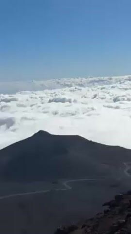 The video shows a view of a dark, volcanic landscape with a caldera in the foreground. A road winds through the dark terrain. Above the volcanic landscape, a layer of white clouds stretches out to the horizon, topped by a clear blue sky. The camera pans slightly to the left.