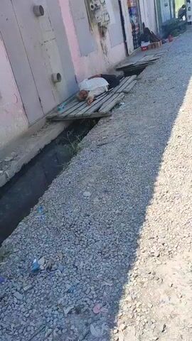 The media file depicts an outdoor scene, likely in Tursunzoda, Tajikistan, judging by the context provided. The central subject is an individual lying on a makeshift wooden platform that spans over a shallow, dark trench. The person is dressed in a light-colored shirt and dark trousers. A bottle of water is visible near the person's head. The platform appears weathered and assembled from salvaged wood, resting against a building with a pale pink and beige facade. The ground is covered in coarse gravel.

The activity taking place is unclear, but the person appears to be resting or possibly unwell, given their supine position and the presence of the water bottle. The lighting suggests it is daytime, with sunlight casting shadows, indicating fair weather. The setting appears to be a somewhat unkempt urban or semi-urban environment, with the building's exterior showing some wear and tear, including visible pipes and what might be an air conditioning unit. The trench appears to be a drainage channel. No text is clearly visible in the frame. The overall impression is one of a person in a vulnerable state in a public or semi-public space.