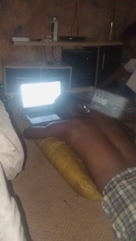The video shows a room with two people and several electronic devices. In the foreground, a shirtless person wearing plaid shorts is lying face down on a bed with a yellow pillow under their stomach. A laptop with a brightly lit screen is positioned in front of them. Another person wearing a white shirt is sitting nearby. The room also contains a television mounted on the wall, a shelf with various items including a bottle, and additional electronic equipment. The walls have a textured, mottled appearance. The video is somewhat blurry and the camera moves around. The sound of a sneeze is heard.