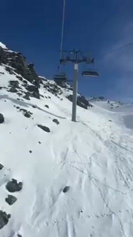 A video depicts a ski lift ascending a snow-covered mountain under a clear blue sky. The lift passes over a slope marked with ski tracks. Inside the lift are two people wearing ski helmets and goggles. The person closest to the camera has iridescent goggles and a grey and white patterned jacket. The second person is wearing a black and white checkered shirt.