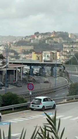 The video shows a highway toll booth in Italy on a cloudy day. There are several cars going through the toll booth. In the foreground, a light blue Renault Twingo is parked on the side of the road near a no-parking sign. The background is a hilly cityscape with buildings of various colors. A few plants are visible in the lower part of the frame.  The camera angle is slightly elevated, as if taken from a nearby building.