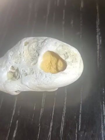 A close-up video shows a light-colored rock with a yellow-orange cavity containing granular material. The rock is placed on a dark, textured surface with visible vertical lines. The camera pans and zooms slightly, offering different perspectives of the object. The lighting creates a glowing effect around the rock.