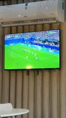 The video shows a large flat-screen television mounted on a wall, displaying a live soccer match. An air conditioning unit is mounted above the television. The wall behind the television and air conditioner is decorated with vertical wooden panels. In the foreground, a portion of a white round table and a white chair are visible. The soccer game features players in white and blue uniforms on a green field, with spectators in the stands. The score and game time are displayed on a graphic overlay in the upper left corner of the screen. The air conditioner displays a temperature of 24 degrees Celsius.