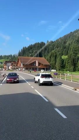 The video shows a road in a mountainous region with trees on the right and buildings on the left. Several cars are driving on the road. A white SUV is in the foreground on the right, and a dark minivan is in the middle of the road. A burgundy car is in the left lane. There is a pedestrian crossing marked on the road. A flag is visible on a pole on the right side of the road. The sky is blue with some clouds.