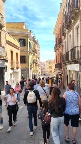 The video shows a street in Cagliari, Italy. People are walking along the street which is lined with buildings. The buildings are a variety of colors, including yellow, orange, and pink. The sky is blue with some clouds. There is a pharmacy on the right side of the street.