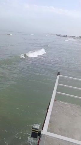 The video begins with a close-up of a pier, showing its concrete surface and white railing. The camera then pans out to reveal the ocean with gentle waves. In the distance, there is a beach lined with trees and buildings, including a distinctive sail-shaped hotel. The camera continues to pan along the coastline, showing more of the ocean, beach, and the pier. The scene is peaceful and serene, with the sound of the waves providing a calming atmosphere. The video ends with a shot of the ocean stretching out to the horizon.