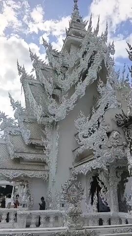 The video shows the exterior of a highly ornate white building, possibly a temple, with intricate, detailed carvings covering its facade and roof. The sky above is partly cloudy and blue. In the foreground, several people are visible. On the left, a person with a red top is standing, and to their right, a person in a dark shirt is looking down. Further to the right and closer to the camera, a woman with dark hair is looking towards the viewer and smiling. Another person is partially visible in the background. The architectural style is elaborate and appears to be of Southeast Asian origin, characterized by pointed spires and flowing decorative elements. The overall impression is one of a visually stunning and culturally significant structure.