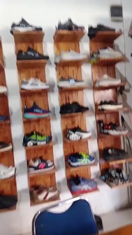 The video displays an indoor setting, likely a shoe store or a showroom, located in Bireuenmeunasahcapa, Indonesia. Numerous pairs of athletic and casual shoes are neatly organized and showcased on vertically stacked wooden shelves attached to a white wall.

The shelves are made of light-colored wood, giving a rustic appearance. Each shelf holds one to two pairs of shoes. A wide variety of shoe brands and styles are visible, including sneakers and running shoes. Recognizable brands include Adidas, Nike, Hoka, New Balance, and Asics, among others. Some shoes appear new, while others show slight signs of wear.

In the foreground, a black oscillating fan is positioned on the floor to the left, and parts of two blue chairs are visible. The room is well-lit, providing clear visibility of all the shoes on display. There are no people visible in the video, and no specific activity is taking place other than the panning shot of the shoe display.